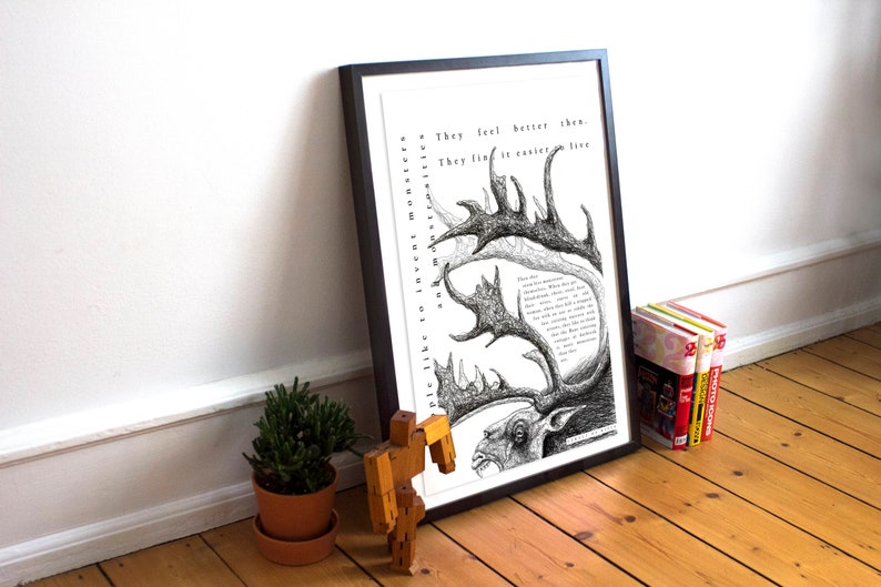 May include: Black framed art print with a detailed pen and ink illustration of a monster with antlers and text. The artwork is propped against a white wall on a wooden floor, with a small potted plant and books nearby.