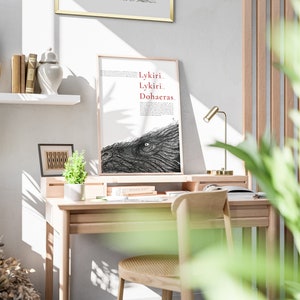 Pu&ograve; includere: Un'illustrazione in bianco e nero incorniciata di un occhio di un grande animale con il testo "Lykiri Lykiri Dohaeras" in rosso. L'illustrazione &egrave; su sfondo bianco ed &egrave; incorniciata in una cornice di legno chiaro. La cornice &egrave; appoggiata su una scrivania di legno con una lampada e una pianta.