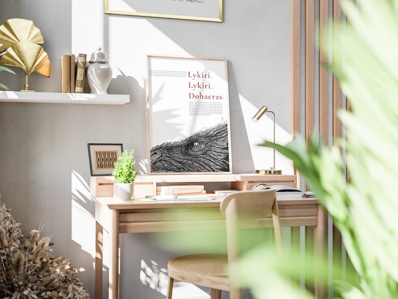 Pu&ograve; includere: Un'illustrazione in bianco e nero incorniciata di un occhio di un grande animale con il testo "Lykiri Lykiri Dohaeras" in rosso. L'illustrazione &egrave; su sfondo bianco ed &egrave; incorniciata in una cornice di legno chiaro. La cornice &egrave; appoggiata su una scrivania di legno con una lampada e una pianta.