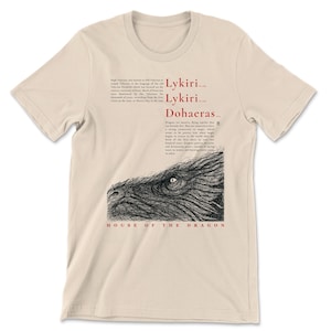 May include: A beige t-shirt with a black and white dragon illustration. The design includes the words "Lykiri, Lykiri, Dohaerus" and "House of the Dragon" in red and black text. The shirt is a neutral colour.