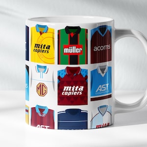 May include: White ceramic mug decorated with a collage of football shirts in various colours. The shirts feature sponsor logos such as "mita copiers" and "müller". The mug has a white handle on the right side.