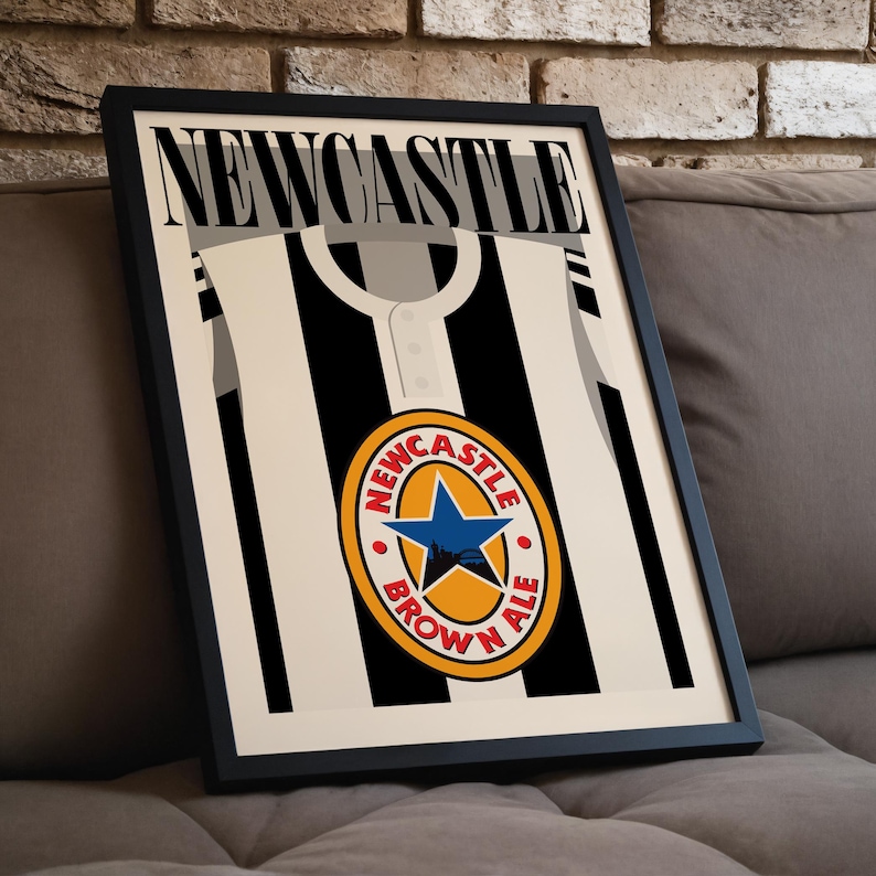 Op de afbeelding: Een zwart-wit ingelijste poster met het woord "NEWCASTLE" in grote witte letters bovenaan. De poster toont een zwart-wit gestreept voetbaltenue met een gele en oranje cirkel in het midden. De cirkel heeft de woorden "NEWCASTLE BROWN ALE" rond de rand en een blauwe ster in het midden.