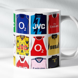 May include: White ceramic mug with a collage of different soccer jerseys. The jerseys feature various sponsors, including JVC, O2, Fly Emirates, and Sega.