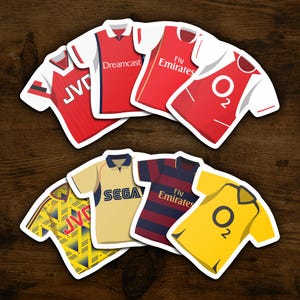May include: A collection of nine different Arsenal football jerseys, featuring various sponsors like JVC, Dreamcast, Fly Emirates, and O2. The jerseys are in red, yellow, blue, and white.