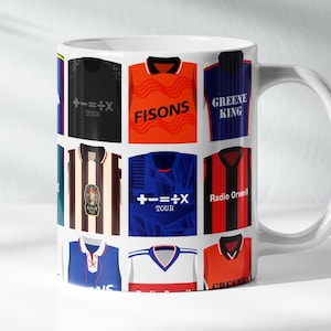 May include: White ceramic mug decorated with a collection of football shirts in various colours. The shirts feature logos and text, including "FISONS" and "Radio Orwell". The mug has a white handle and is suitable for hot beverages.