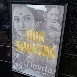 May include: A black and white movie poster for the film "Devdas" starring Aishwarya Rai, Madhuri Dixit, and Shah Rukh Khan. The poster features the text "NOW SHOWING" in yellow neon letters.