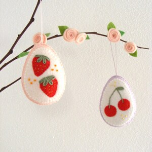 May include: Two felt ornaments hanging from a branch. One ornament is pink with two embroidered strawberries. The other ornament is purple with two embroidered cherries.