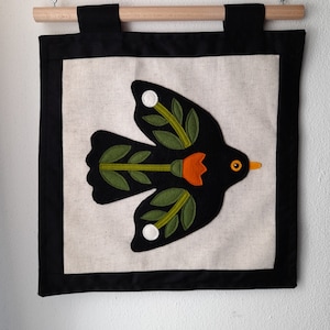 Op de afbeelding: Een zwarte en groene vogel met oranje accenten is geborduurd op een beige vierkant van stof. Het vierkant is omlijst met een zwarte rand en hangt aan een houten stok met een touw.