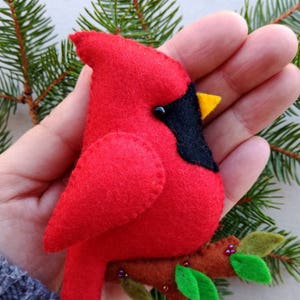 May include: A felt cardinal ornament with a red body, black head, and yellow beak. The cardinal is perched on a brown felt branch with green felt leaves.