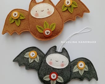 May include: Two felt bat ornaments with embroidered details. One bat is brown with orange and yellow flowers, the other is gray with orange and yellow flowers. Both bats have white faces with pink cheeks and a stitched smile. The bats are hanging from white strings. The text "NUVOLINA HANDMADE" is visible in the image.