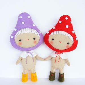 May include: Two felt mushroom dolls holding hands. The doll on the left has a purple mushroom hat with white polka dots and yellow boots. The doll on the right has a red mushroom hat with white polka dots and green boots.