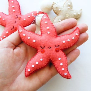 May include: Two red felt starfish with white bead accents and red button eyes. The starfish are held in a hand.