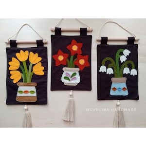 Può includere: Tre arazzi da parete in feltro con motivi floreali. Ogni arazzo presenta un fiore diverso in un vaso: tulipani gialli, fiori rossi e fiori bianchi a forma di campana. Gli arazzi sono su uno sfondo marrone scuro e hanno un'asta di legno in alto con un fiocco bianco.