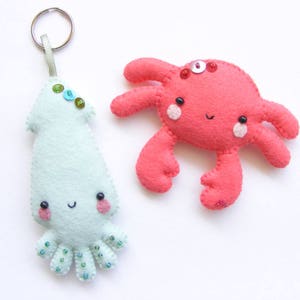 May include: A set of two felt animal keychains. The first keychain is a light green squid with a smiling face and colorful beads. The second keychain is a red crab with a smiling face and a single pink bead on its head.