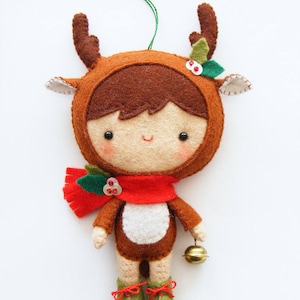 May include: A felt reindeer ornament with a brown and white reindeer costume, red scarf, green holly, and a gold bell. The reindeer has a smiling face with brown hair and black eyes.