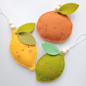 May include: Three felt fruit decorations, a lemon, an orange, and a lime, with smiling faces and green felt leaves. Each decoration has a white string for hanging.