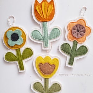 Op de afbeelding: Vier viltbloemen ornamenten met groene bladeren en stelen. De bloemen zijn geel, oranje en bruin. De ornamenten hangen aan een wit touwtje.
