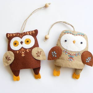 May include: Two felt owl ornaments with embroidered details. One owl is brown with yellow eyes and the other is beige with white eyes. Both owls have brown wings and are hanging from a string.