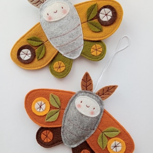 Op de afbeelding: Twee vilten motornamenten. Elke mot heeft een grijze body met een lachend gezicht en decoratieve vleugels in tinten oranje, geel, bruin en groen. De ornamenten hebben een touwtje om op te hangen.