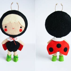 Puede incluir: Una muñeca de fieltro con forma de mariquita con alas negras y rojas, detalles blancos y zapatos verdes. La muñeca tiene una cabeza negra con una cara blanca y cabello rubio.