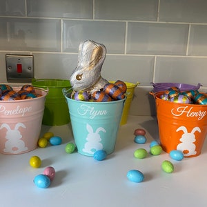 May include: Three pastel-coloured metal buckets filled with chocolate Easter eggs. The buckets are labelled "Penelope", "Flynn", and "Henry". Each bucket has a white bunny silhouette on the front.