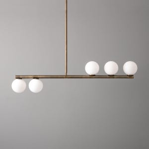 May include: A modern brass and glass chandelier with five white globe-shaped lights. The chandelier has a long, horizontal bar with the lights evenly spaced along it. The bar is connected to a vertical rod that hangs from the ceiling.