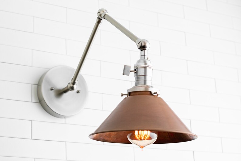 May include: A wall-mounted, adjustable lamp with a brushed nickel finish. The lamp features a conical, copper-colored shade and an exposed Edison-style bulb. The fixture is mounted on a white tiled wall, showcasing a modern, industrial design.