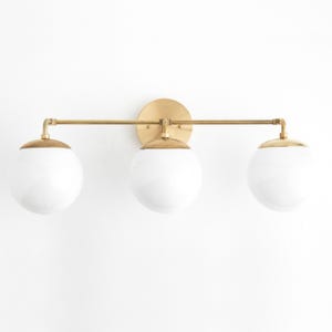 May include: A three-light vanity fixture with a brass finish. The fixture features three spherical, white glass shades. The light is mounted on a round brass backplate and has a horizontal bar.