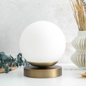 May include: A table lamp featuring a spherical white glass shade resting on a circular brushed brass base. The lamp has a smooth, matte metallic finish on the bottom stand and a clean, opaque white globe top that provides a simple, rounded form.