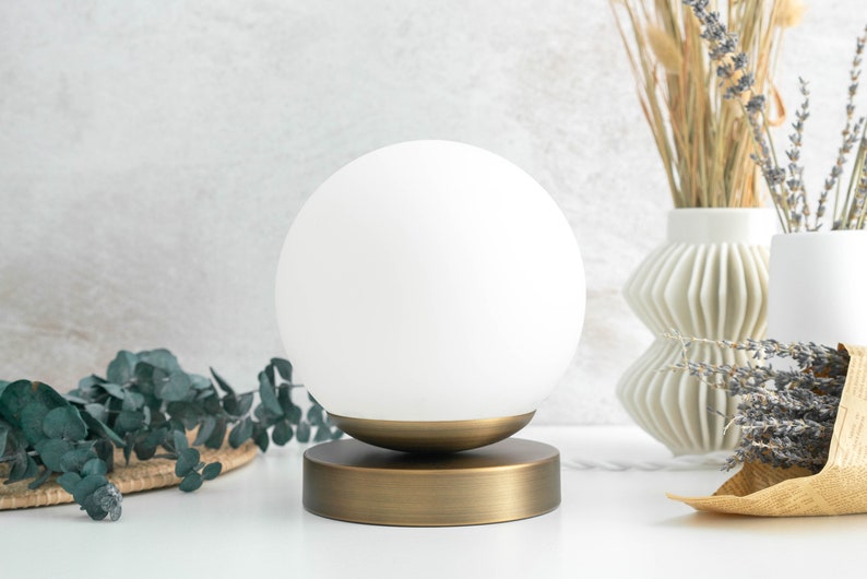 May include: A table lamp featuring a spherical white glass shade resting on a circular brushed brass base. The lamp has a smooth, matte metallic finish on the bottom stand and a clean, opaque white globe top that provides a simple, rounded form.