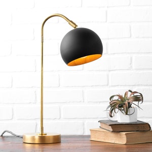 May include: A black and gold desk lamp with a round shade. The lamp is on a wooden table in front of a white brick wall. A small plant is sitting on a stack of books.