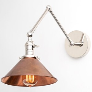 May include: A wall-mounted, adjustable lamp with a copper-colored conical shade and a silver-toned, articulated arm. The lamp features a visible lightbulb and a circular mounting plate. The design is industrial and modern.