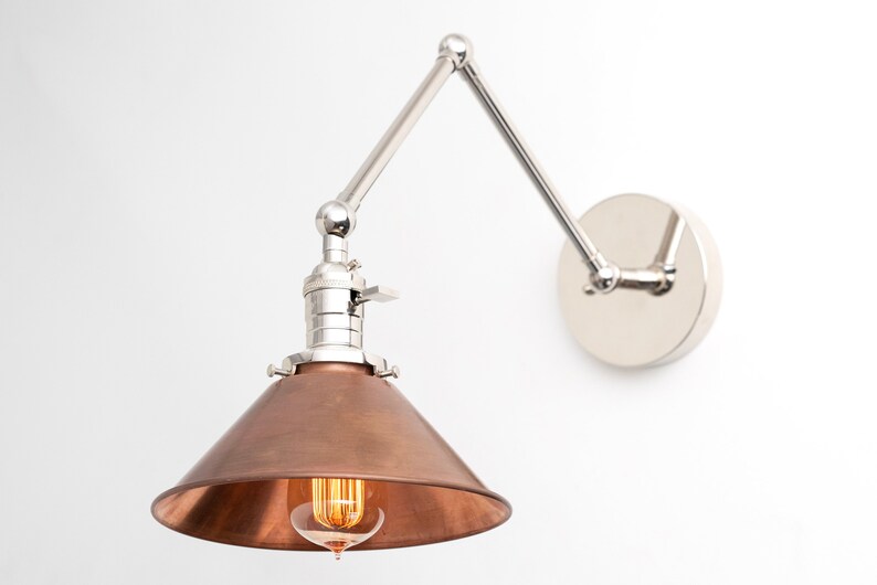 May include: A wall-mounted, adjustable lamp with a copper-colored conical shade and a silver-toned, articulated arm. The lamp features a visible lightbulb and a circular mounting plate. The design is industrial and modern.