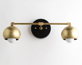 May include: A modern wall sconce with two spherical, gold-colored light fixtures. The lights are attached to a brass-colored bar and a black circular base. Each light fixture has a white bulb.