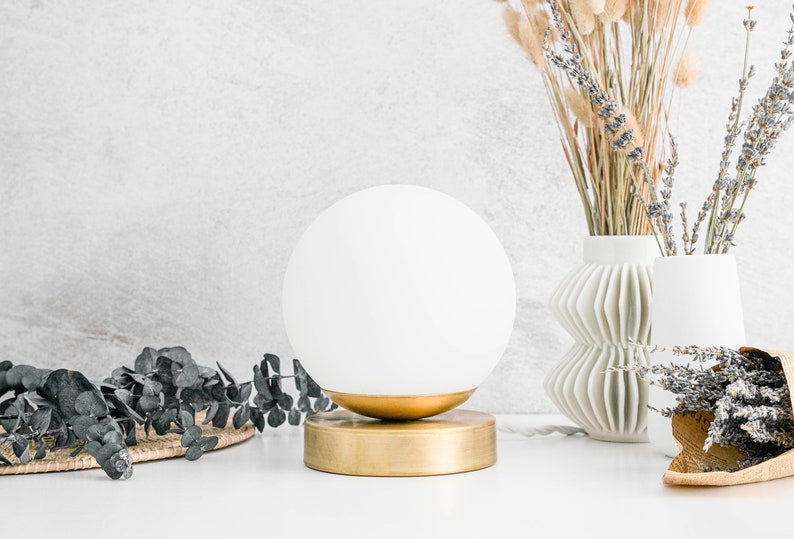 May include: A spherical white glass lamp shade sits atop a circular brushed gold metal base. The lamp features a smooth, opaque white globe that rests securely in a gold-toned metal cup, which is attached to a flat, cylindrical gold metal stand.