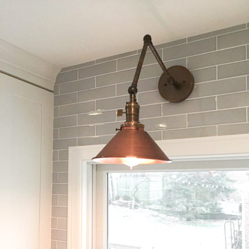 May include: A vintage-style wall-mounted lamp with a copper-colored shade and adjustable arm. The lamp features a brass-colored base and a round mounting plate. It is installed on a gray brick-patterned wall, near a window.