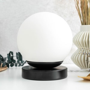 May include: A spherical white glass lamp shade sits atop a circular black metal base. The lamp features a smooth, opaque white globe that rests securely on the matte black cylindrical stand, creating a clean and balanced lighting fixture design.