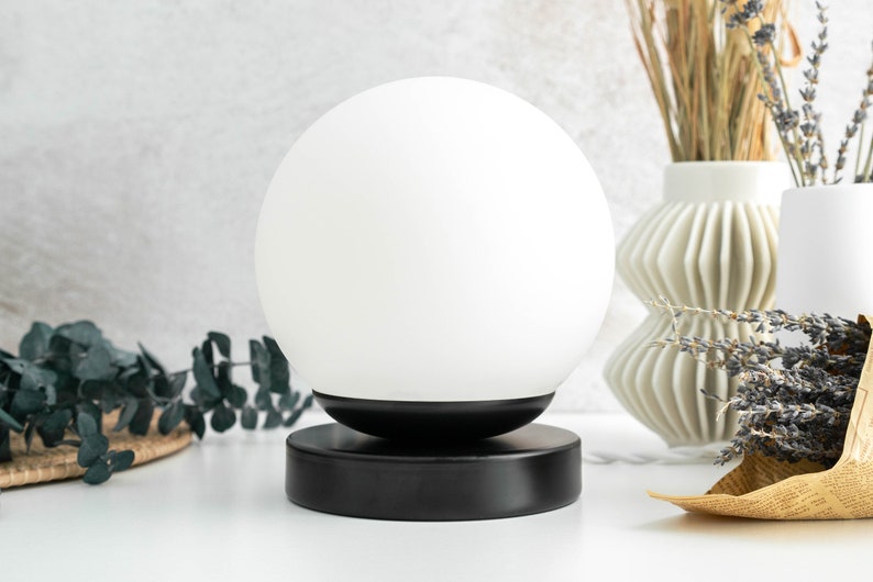 May include: A spherical white glass lamp shade sits atop a circular black metal base. The lamp features a smooth, opaque white globe that rests securely on the matte black cylindrical stand, creating a clean and balanced lighting fixture design.