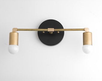 May include: A wall-mounted light fixture featuring a circular matte black base plate and a horizontal brass-toned metal bar. Two cylindrical brass-toned sockets are attached to the ends, each holding a round white glass light bulb.