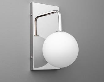 May include: A modern wall sconce with a polished chrome finish. It features a rectangular backplate, a curved arm, and a spherical white glass shade. The sconce is mounted on a gray wall, providing a clean, contemporary look.