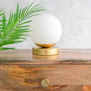 May include: A table lamp featuring a spherical white frosted glass shade resting on a circular brushed gold metal base. The lamp includes a white twisted fabric power cord extending from the side of the base, providing a clean and simple design.