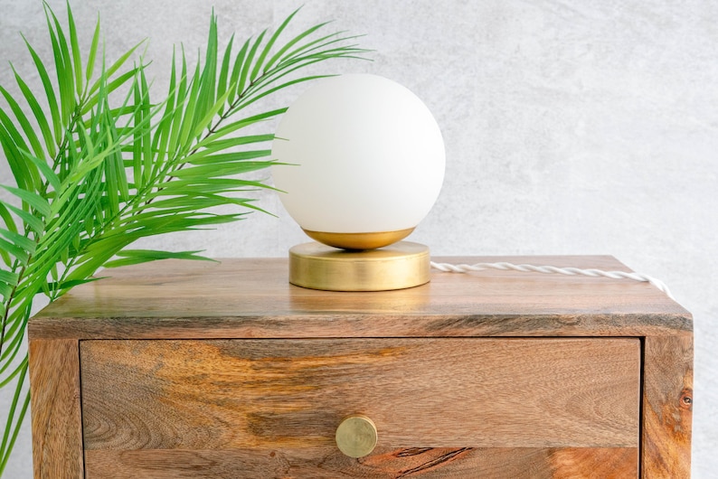 May include: A table lamp featuring a spherical white frosted glass shade resting on a circular brushed gold metal base. The lamp includes a white twisted fabric power cord extending from the side of the base, providing a clean and simple design.