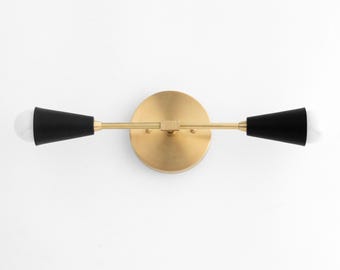 May include: A gold wall sconce with two black cone-shaped shades. The sconce has a round base and a straight arm.