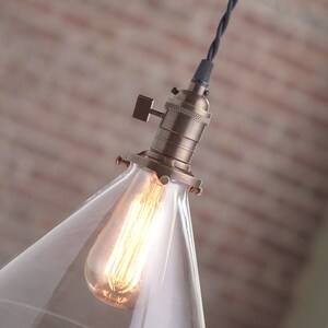 May include: A clear glass cone pendant light fixture with a brass socket and a black wire. The light bulb is turned on and glowing.