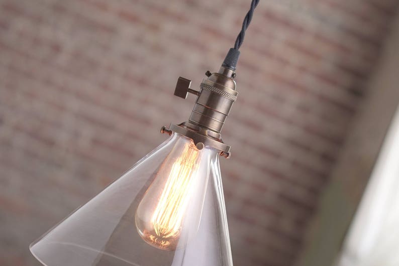 May include: A clear glass cone pendant light fixture with a brass socket and a black wire. The light bulb is turned on and glowing.