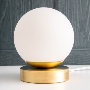 May include: A table lamp featuring a spherical white frosted glass shade resting on a circular brushed gold metal base. The lamp includes a white power cord extending from the side of the base, providing a clean and simple lighting fixture design.