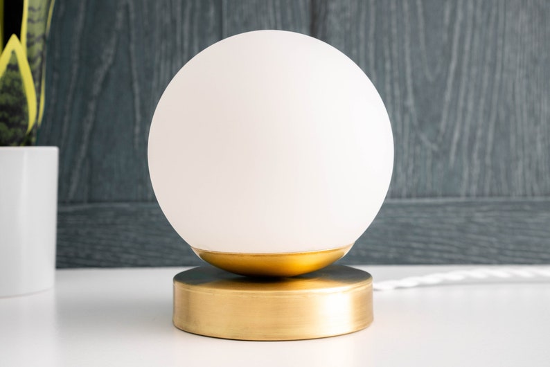 May include: A table lamp featuring a spherical white frosted glass shade resting on a circular brushed gold metal base. The lamp includes a white power cord extending from the side of the base, providing a clean and simple lighting fixture design.