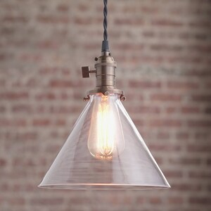 May include: A clear glass pendant light with a brass finish and a cone-shaped shade. The light is hanging from a black cord.