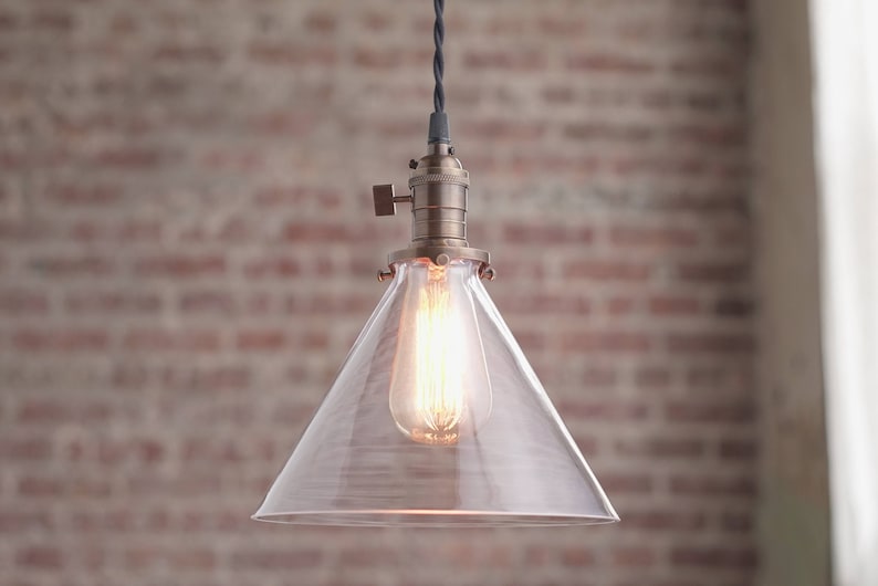May include: A clear glass pendant light with a brass finish and a cone-shaped shade. The light is hanging from a black cord.