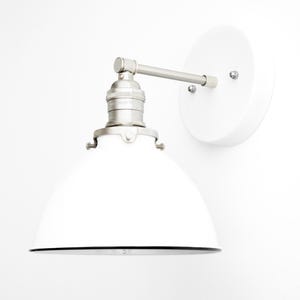 Puede incluir: Un aplique de pared con una pantalla de metal blanca en forma de c&uacute;pula, un casquillo de n&iacute;quel cepillado y una placa de pared circular blanca a juego. El brazo conecta la pantalla a la base con una bisagra visible.